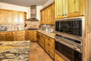 Kitchen featuring appliances with stainless steel finishes, wall chimney exhaust hood, tasteful backsplash, light stone countertops, and light tile patterned floors