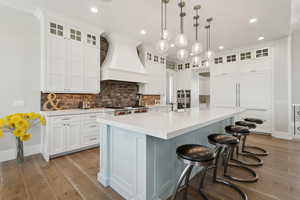 Kitchen featuring custom exhaust hood, wood finished floors, light countertops, a spacious island, and decorative backsplash