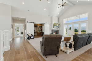 Living area featuring light wood finished floors, a ceiling fan, beam ceiling, a barn door, and high vaulted ceiling