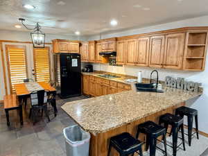 Kitchen featuring black refrigerator with ice dispenser, a peninsula, gas cooktop, under cabinet range hood, and light stone counters. Doors exit to the back patio.