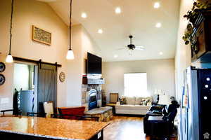 Living area with a stone fireplace, a barn door, ceiling fan, light wood-type flooring, and high vaulted ceiling