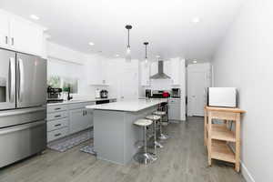 Kitchen featuring stainless steel appliances, wall chimney exhaust hood, a kitchen bar, light wood-type flooring, and recessed lighting