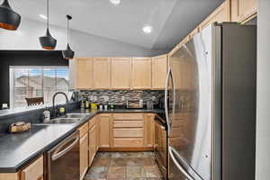 Kitchen featuring stainless steel appliances, light brown cabinetry, lofted ceiling, dark countertops, and decorative backsplash