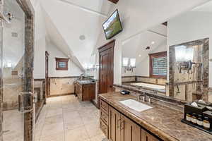 Bathroom featuring vaulted ceiling, two vanities, a shower stall, a garden tub, and a wainscoted wall