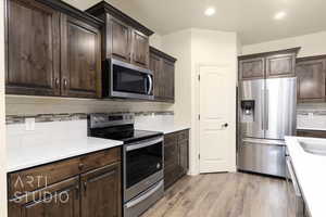 Kitchen with stainless steel appliances, dark brown cabinets, backsplash, wood finished floors, and light countertops