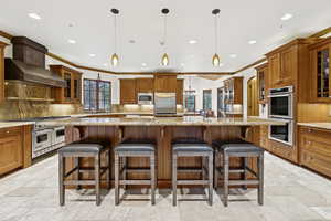 Kitchen featuring stainless steel appliances, brown cabinets, crown molding, stone tile flooring, and wall chimney range hood