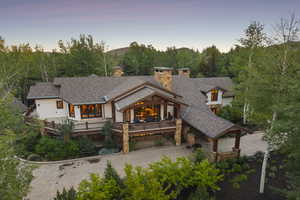 Back of house at dusk with a chimney, driveway, stone siding, a wooded view, and a shingled roof