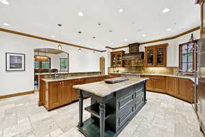 Kitchen with premium range hood, arched walkways, a large island with sink, brown cabinetry, and ornamental molding