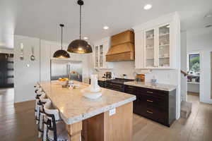Kitchen featuring premium appliances, custom exhaust hood, light wood finished floors, recessed lighting, and light stone counters