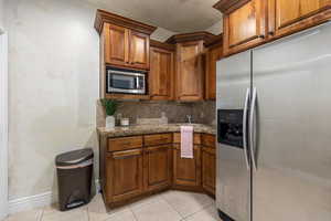 Kitchen featuring appliances with stainless steel finishes, backsplash, dark stone counters, brown cabinetry, and light tile patterned floors