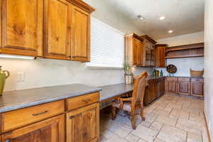 Kitchen with wood finish cabinetry, stone tile flooring, recessed lighting, built in study area, and glass fronted cabinets