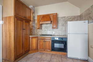 Kitchen with freestanding refrigerator, black oven, custom exhaust hood, brown cabinetry, and vaulted ceiling