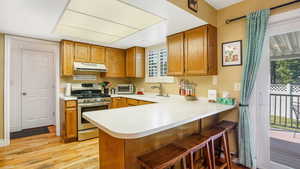Kitchen with stainless steel range with gas stovetop, under cabinet range hood, light wood-type flooring, light countertops, and brown cabinets