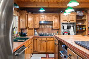 Kitchen with tile counters, appliances with stainless steel finishes, a wood ceiling with exposed beams, under cabinet range hood, and tasteful backsplash