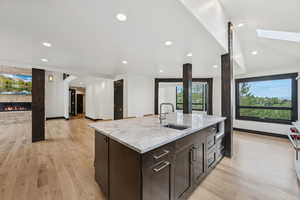 Kitchen featuring a stone fireplace, light wood-style floors, a kitchen island with sink, dark brown cabinets, and recessed lighting