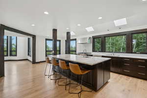 Kitchen featuring wall chimney range hood, a skylight, a breakfast bar, a center island with sink, and light stone countertops