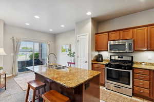Kitchen featuring appliances with stainless steel finishes, light stone counters, brown cabinetry, light colored carpet, and an island with sink