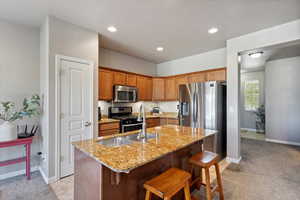 Kitchen featuring stainless steel appliances, an island with sink, a breakfast bar, light carpet, and light stone countertops