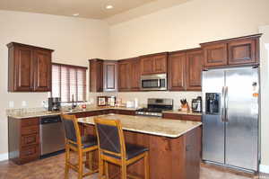 Kitchen with appliances with stainless steel finishes, high vaulted ceiling, light stone countertops, a kitchen breakfast bar, and a center island