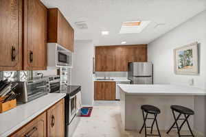 Kitchen featuring stainless steel appliances, brown cabinetry, light floors, a breakfast bar area, and light countertops