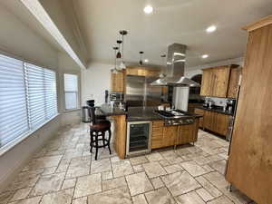Kitchen featuring beverage cooler, island range hood, appliances with stainless steel finishes, crown molding, and stone tile flooring