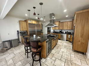 Kitchen featuring island range hood, appliances with stainless steel finishes, beverage cooler, stone tile floors, and a center island with sink