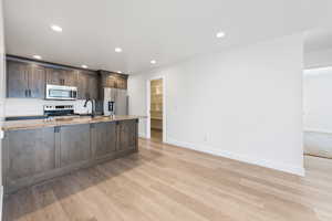 Kitchen featuring appliances with stainless steel finishes, light wood-style floors, dark brown cabinetry, recessed lighting, and light stone counters