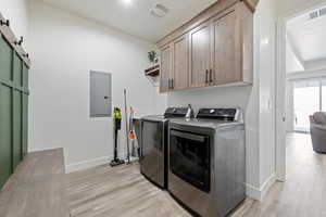 Laundry area with electric panel, washer and dryer, cabinet space, light wood-style floors, and a barn door