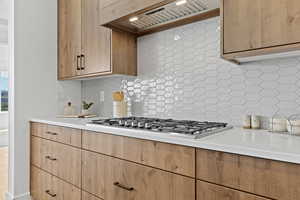 Kitchen featuring decorative backsplash, custom exhaust hood, stainless steel gas stovetop, light brown cabinetry, and light stone counters