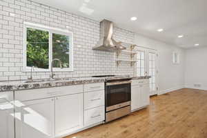 Kitchen with electric range, wall chimney exhaust hood, tasteful backsplash, light stone counters, and white cabinetry
