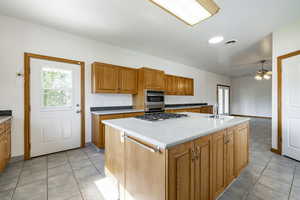 Kitchen with stainless steel appliances, ceiling fan, brown cabinetry, light tile patterned flooring, and an island with sink