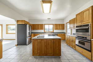 Kitchen featuring stainless steel appliances, an island with sink, brown cabinets, light tile patterned floors, and light countertops