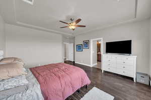 Bedroom with a tray ceiling, dark wood-style floors, ceiling fan, and ensuite bath
