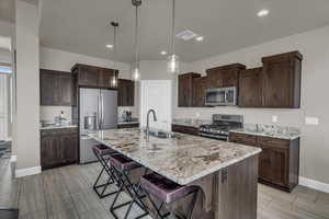 Kitchen featuring appliances with stainless steel finishes, a breakfast bar area, dark brown cabinets, decorative light fixtures, and light stone countertops
