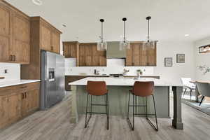Kitchen featuring brown cabinetry, an island with sink, stainless steel refrigerator with ice dispenser, pendant lighting, and tasteful backsplash