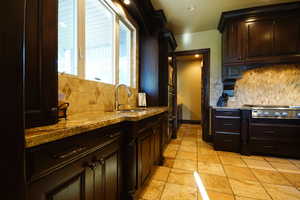 Kitchen featuring stainless steel gas cooktop, tasteful backsplash, light stone counters, stone tile floors, and dark brown cabinets