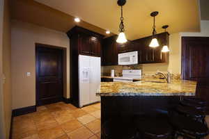 Kitchen with white appliances, a peninsula, light stone counters, dark brown cabinetry, and hanging light fixtures