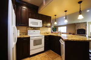 Kitchen with white appliances, a peninsula, a stone fireplace, open floor plan, and decorative light fixtures