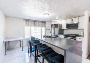 Kitchen featuring dark countertops, stainless steel appliances, white cabinetry, a textured ceiling, and a center island with sink