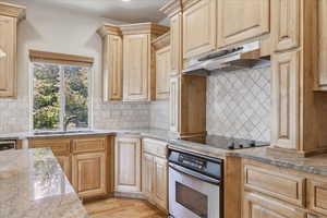 Kitchen featuring stainless steel oven, under cabinet range hood, decorative backsplash, black electric stovetop, and light brown cabinets