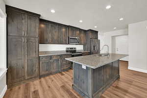 Kitchen featuring stainless steel appliances, light wood-style floors, a kitchen island with sink, dark brown cabinetry, and recessed lighting