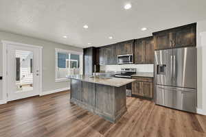 Kitchen with stainless steel appliances, wood finished floors, a kitchen island with sink, a textured ceiling, and light stone countertops