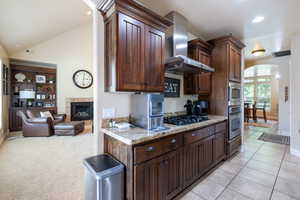 Kitchen featuring arched walkways, appliances with stainless steel finishes, wall chimney exhaust hood, a tile fireplace, and light stone countertops