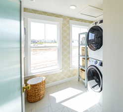 Laundry room featuring stacked washing machine and dryer, light marble finish flooring, wallpapered walls, and attic access
