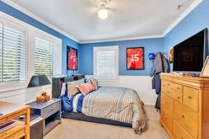 Bedroom featuring multiple windows, a wainscoted wall, light colored carpet, crown molding, and ceiling fan
