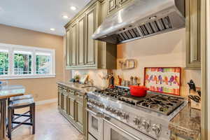 Kitchen with under cabinet range hood, range with two ovens, cream cabinets, light stone counters, and recessed lighting