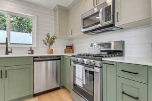 Kitchen featuring green cabinetry, appliances with stainless steel finishes, light countertops, light wood-type flooring, and backsplash