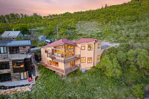 Back of house at dusk featuring a balcony, a forest view, and stone siding