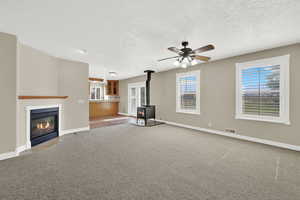 Unfurnished living room with a wood stove, a ceiling fan, a textured ceiling, a fireplace with flush hearth, and carpet floors