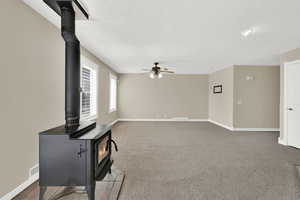 Unfurnished living room featuring a wood stove, a textured ceiling, a ceiling fan, and carpet flooring